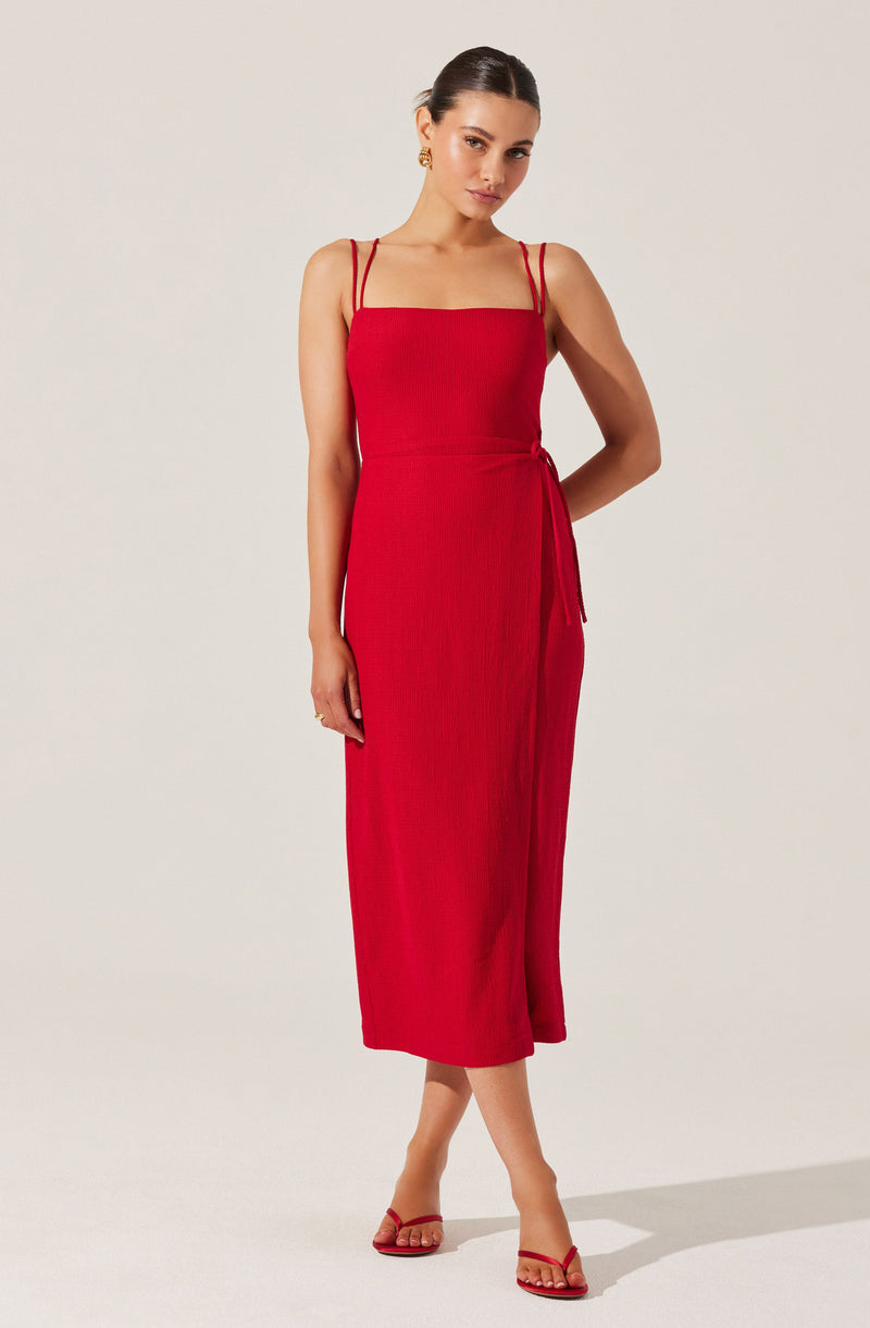 A woman is wearing a sleeveless, red, midi-length dress with thin double straps and a side tie detail. She is accessorized with gold earrings and red strappy high heels.