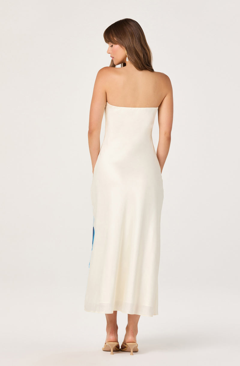 Woman in a strapless, cream-colored, full-length dress with a slit on the side, wearing beige high-heeled sandals.