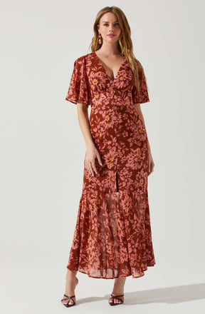 A woman is wearing a long, flowing, floral dress with shades of pink and orange on a dark reddish-brown background. The dress has short, ruffled sleeves and a V-neckline. The fabric features a large floral pattern, and the dress appears semi-sheer with a slit down the front. She is also wearing black strappy high-heeled sandals and statement earrings.