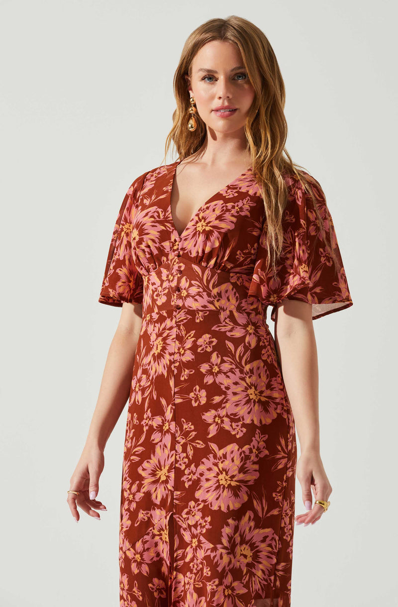A woman wearing a flowing, floral-patterned dress with short, puffed sleeves. The dress has a deep V neckline, and the pattern features large pink and yellow flowers on a dark red background. She is accessorized with gold drop earrings and a gold ring on her right hand.