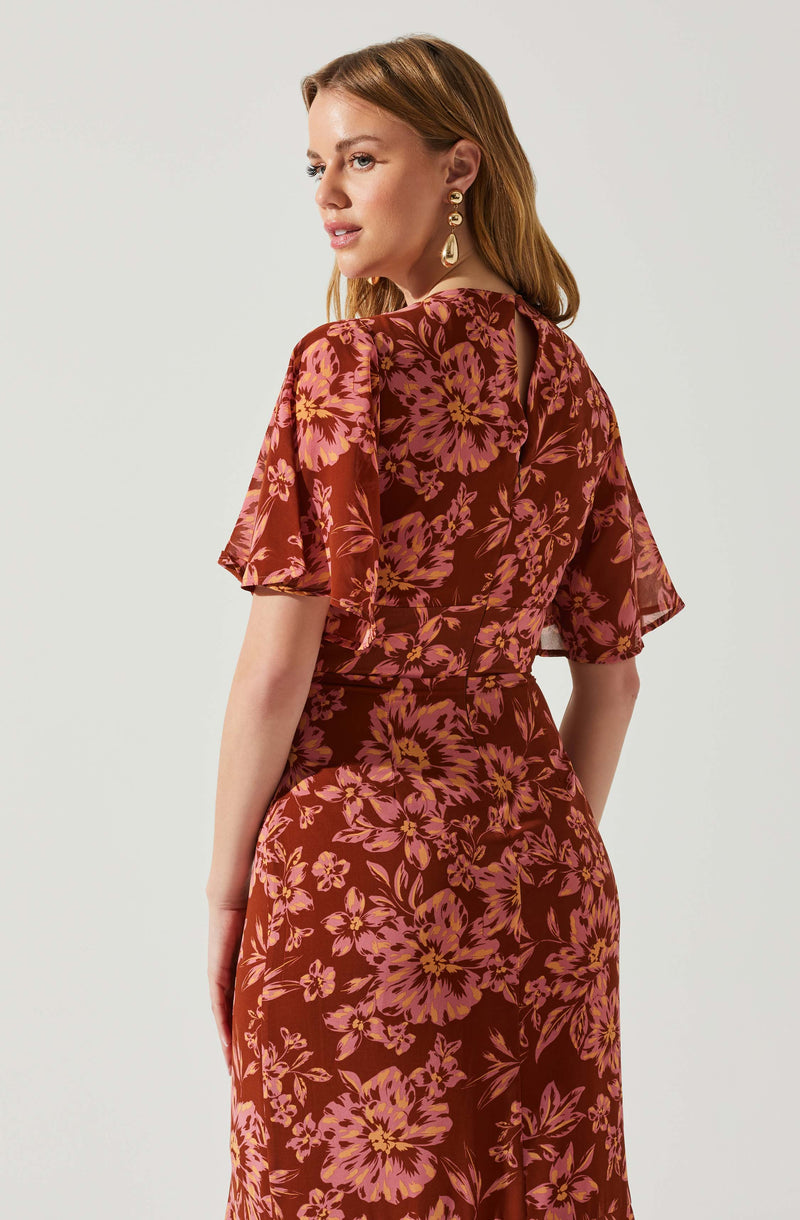 A woman is wearing a matching floral dress with a deep rust background and pinkish-orange flowers. The dress has short, flutter sleeves and a fitted waist. The fabric appears lightweight and flowy. She accessorizes with large, gold, statement earrings.