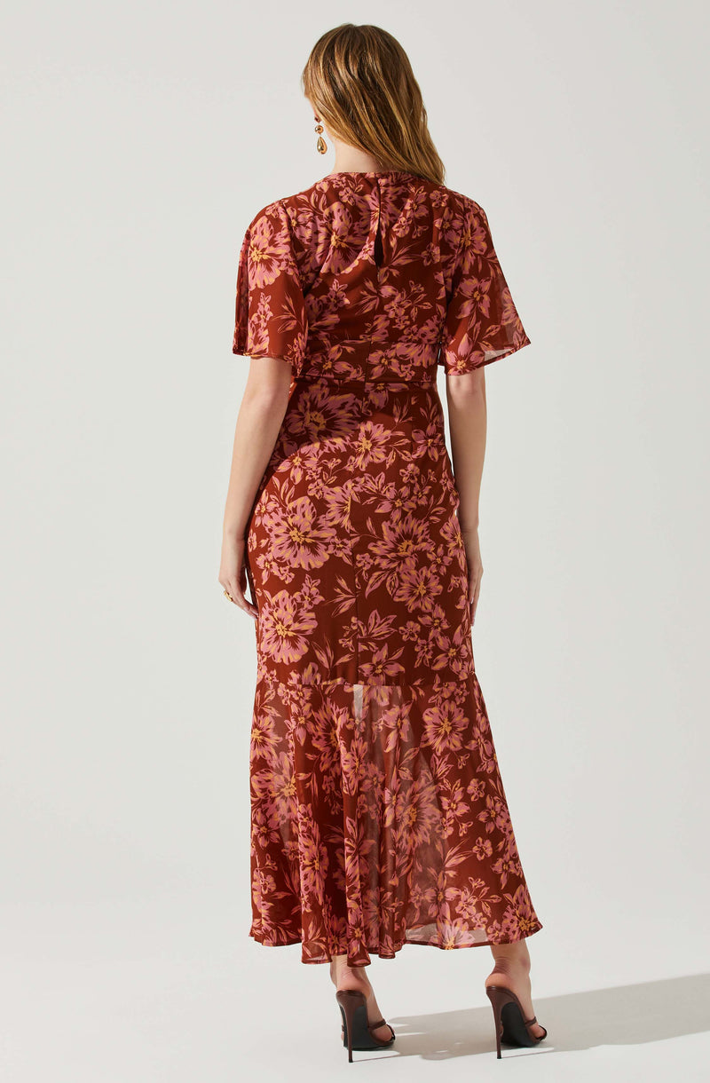 A woman is wearing a long, flowing dress with a floral pattern in shades of pink and purple on a dark red background. The dress features short, puffed sleeves and a fitted waist, with a sheer, ruffled hem at the bottom. She has on high-heeled, open-toed shoes and is accessorized with dangling earrings.