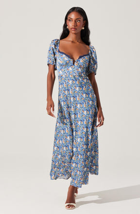 A woman is wearing a long, flowy dress with a blue floral pattern. The dress features puffed short sleeves, a sweetheart neckline with dark lace trim, and a fitted waist that flows into a full-length skirt. She is also wearing open-toe white heels and has long, dark hair styled in soft waves.