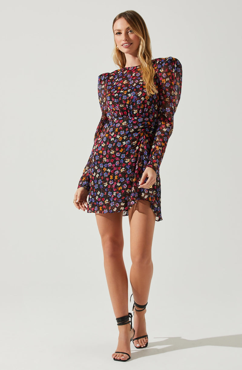 A woman is wearing a black dress with a colorful floral pattern, featuring mostly pink, purple, and blue flowers. The dress has long, sheer sleeves with puffed shoulders and a high neckline. The skirt of the dress is slightly ruffled and falls above the knees. She is also wearing black, strappy high-heeled sandals that wrap around her ankles.