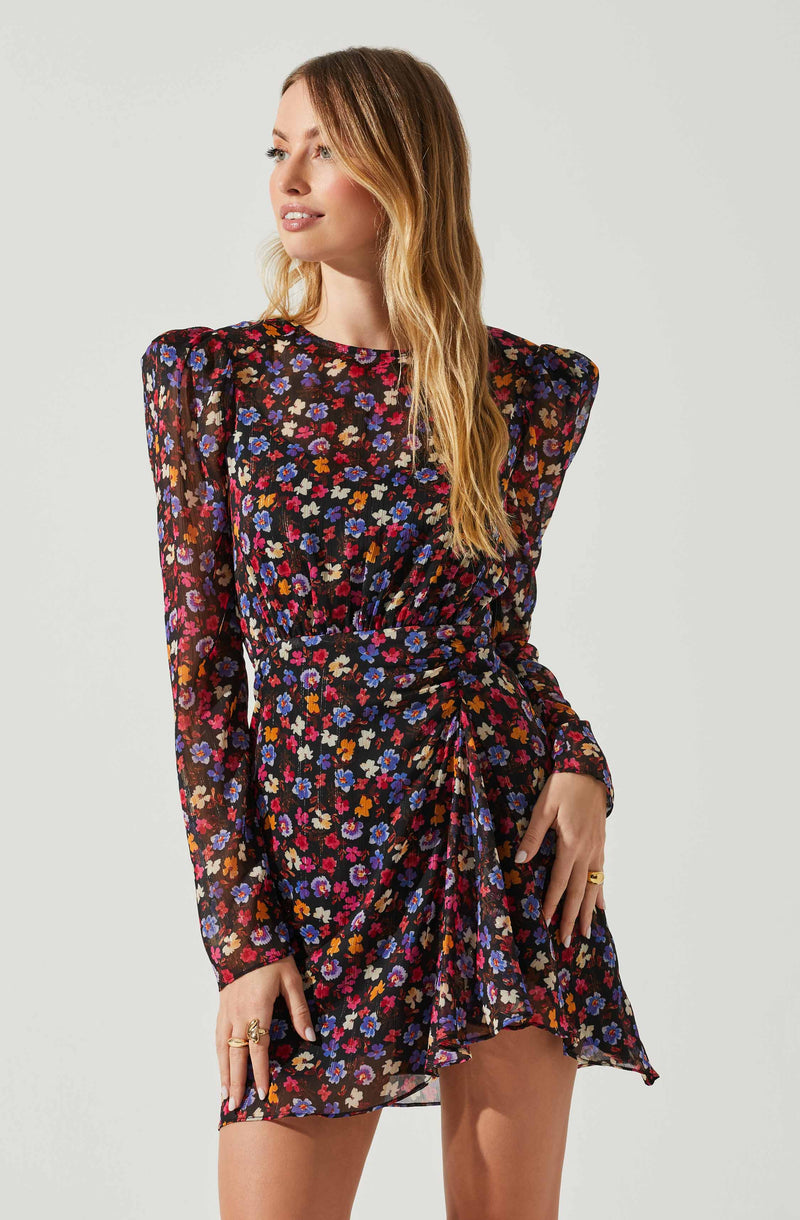 A woman is wearing a long-sleeved, black dress with a colorful floral print featuring shades of blue, red, pink, orange, and white. The dress has puffed shoulders and a gathered, asymmetrical front detail.