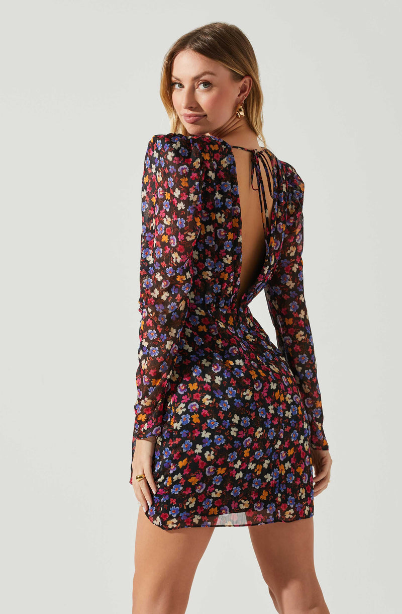 A woman is wearing a black dress with a colorful floral pattern. The dress has long, sheer sleeves, and a deep open back closed with a tie, revealing her upper back.
