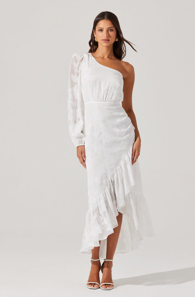 A woman is wearing a white, one-shoulder dress with a ruffled uneven hem that is longer in the back. The dress has a textured pattern and an asymmetrical design, with one long sleeve on her left arm and the right shoulder and arm exposed. She is also wearing white high-heeled sandals.