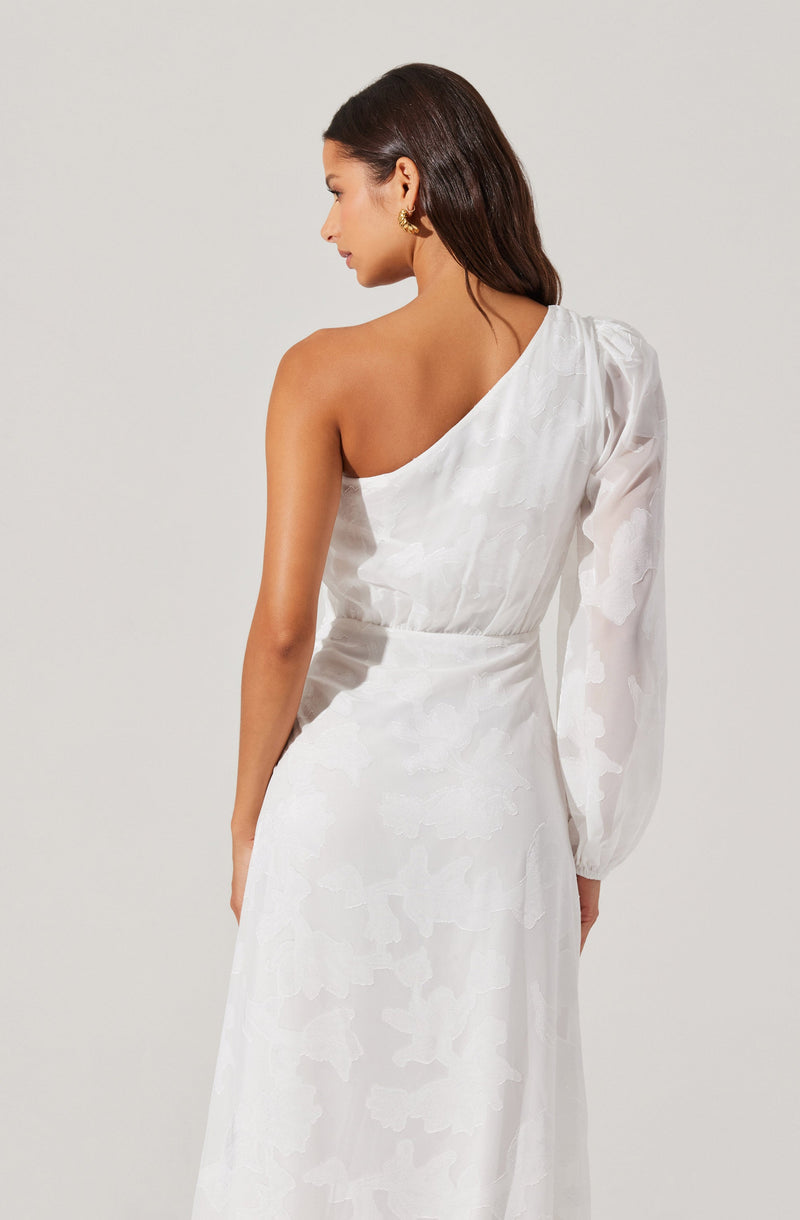 A woman is wearing a white, one-shoulder dress with a sheer, long sleeve on her left arm. The dress features a subtle floral or abstract pattern and a fitted waist. She has dark, wavy hair and is accessorized with gold earrings.