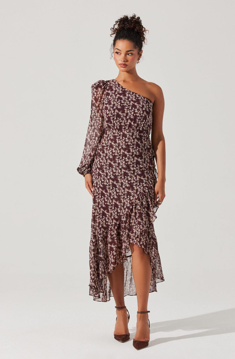 A woman is wearing a long, flowy dress with a high-low hemline. The dress features a dark background with a small, light-colored floral print. It has an asymmetrical, off-the-shoulder neckline, with one long puffed sleeve on the left side and sleeveless on the right. The dress is fitted at the waist and cascades down in ruffled layers, ending above the ankles in the front and extending to the ankles at the back. She is wearing brown high-heeled shoes.