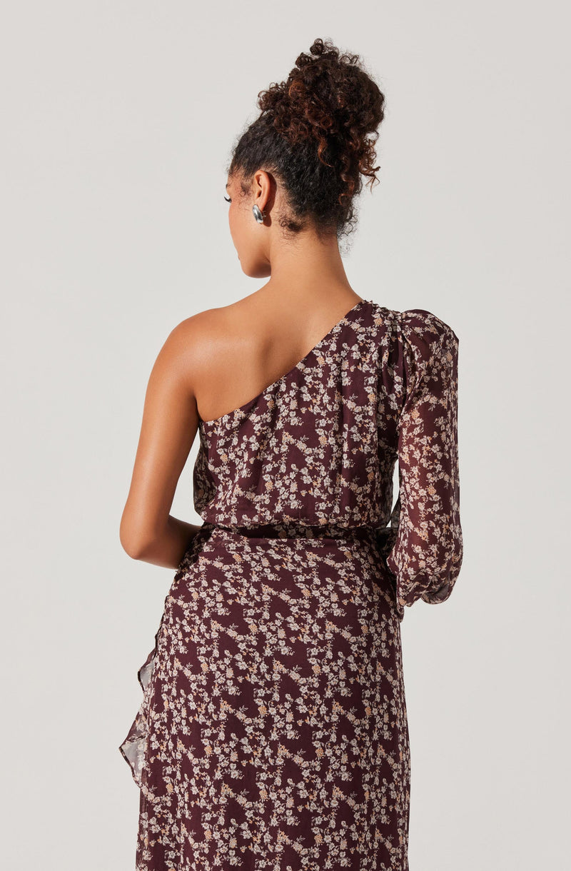 A woman is wearing a one-shoulder dress with a dark background and a floral pattern in light colors. The dress has a single long sleeve on the right side, while the left shoulder is bare. The fabric appears lightweight and flowy.