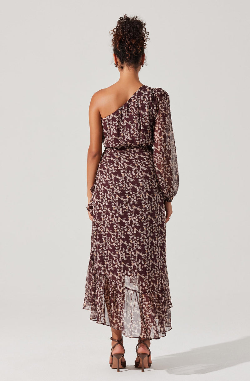 A woman is wearing a long, flowy dress with a dark background and a light floral pattern. The dress features a one-shoulder design with a long, semi-sheer sleeve, and a fitted waist. The skirt of the dress is pleated with a ruffled hem that reaches the floor. She is also wearing high-heeled shoes with ankle straps.