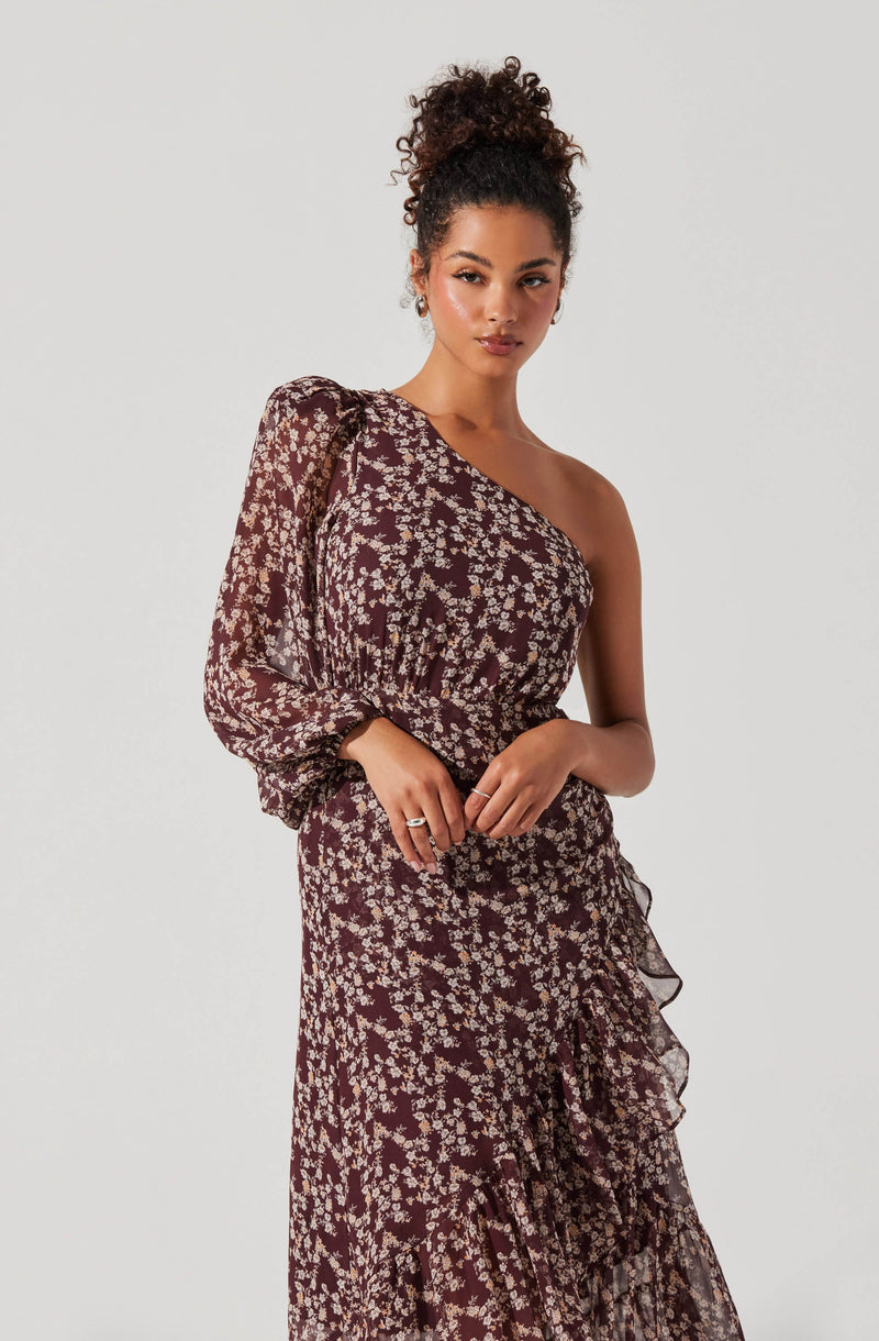 A woman is wearing a dark-colored dress with a floral pattern in light beige or cream. The dress features a one-shoulder design, with a long, sheer sleeve on the left side that is slightly puffed at the shoulder. The fabric of the dress appears lightweight with a ruffled detail running diagonally across the skirt. The overall look is elegant and stylish.