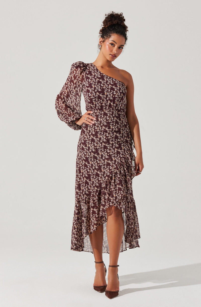 A woman is wearing a dress with a one-shoulder design. The dress has a fitted bodice and a high-low hemline with ruffled detailing. It features a floral pattern in shades of dark red, pink, and cream on a dark background. The dress has long, sheer, puffed sleeves on one side, while the other shoulder remains bare.