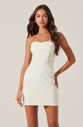 A woman is wearing a strapless, fitted white dress that has a scalloped lace trim along the top edge. The dress features a structured bodice with vertical seam detailing, and the length is above the knees. She accessorizes with gold hoop earrings and has long, wavy brown hair.