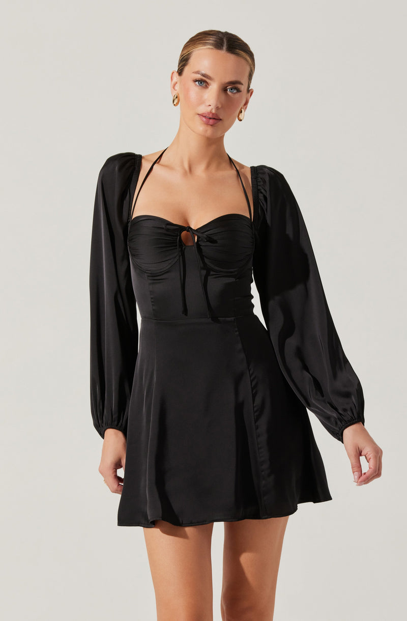 A woman is wearing a black satin dress featuring long, puffed sleeves with elastic cuffs, a fitted bodice with ruched detailing at the bust, and a small keyhole opening with a tie closure at the center of the chest. The dress has a slightly flared skirt that falls above the knees. She accessorizes with gold hoop earrings.