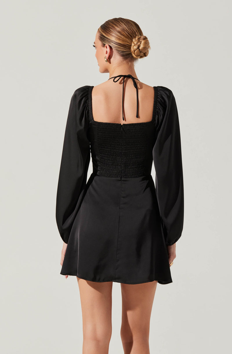 The woman is wearing a black dress with puffy, long sleeves and a fitted, smocked bodice. The dress features a square neckline at the back, with a thin black tie that is tied into a bow at the top. The skirt of the dress is short and flares out slightly.