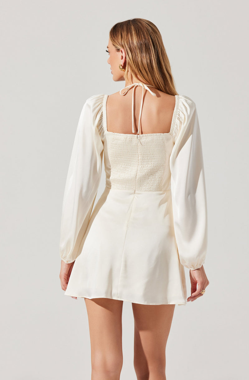 A woman is wearing a cream-colored dress with long puffed sleeves and a square neckline. The dress has a fitted, smocked bodice at the back with a delicate tie strap around the neck. The skirt is short and slightly flared.