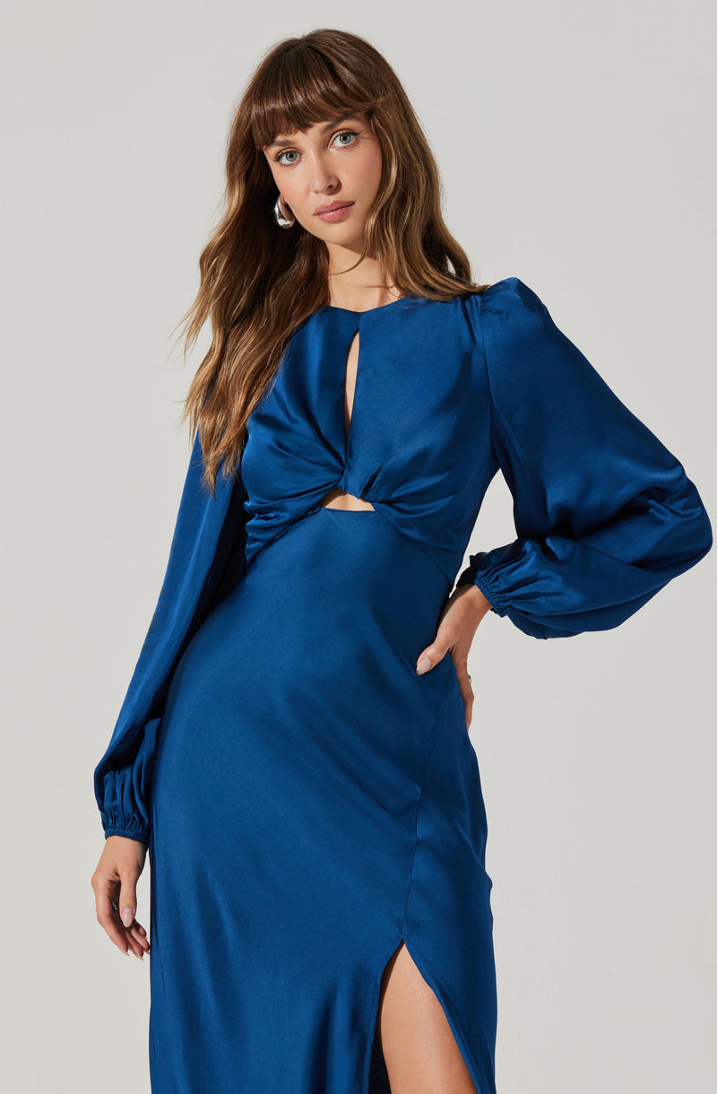 A woman is wearing a long, shiny, royal blue satin dress with long puffed sleeves gathered at the wrists. The dress features a keyhole neckline with a twist knot detail at the bust and a thigh-high slit on one side.