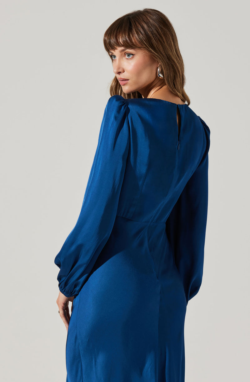A woman is wearing a rich cobalt blue dress made of shiny, satin-like fabric. The dress features long, voluminous sleeves with gathered cuffs and a high neckline. The back of the dress has a small keyhole opening, and the fitted waist accentuates her figure.