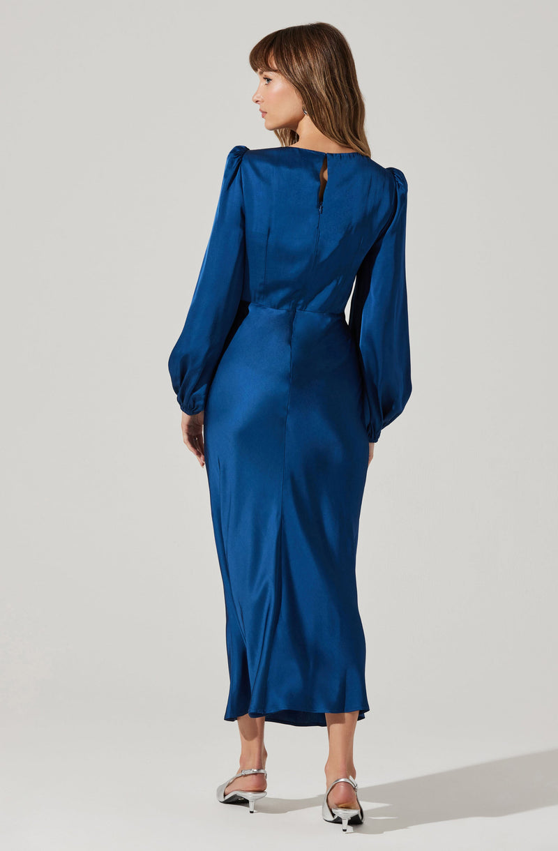 A woman is wearing a long, satin royal blue dress with a fitted waist and a slit at the back. The dress features long, puffed sleeves that gather at the wrists, and a high neckline with a small keyhole opening at the back. She is also wearing white heels.