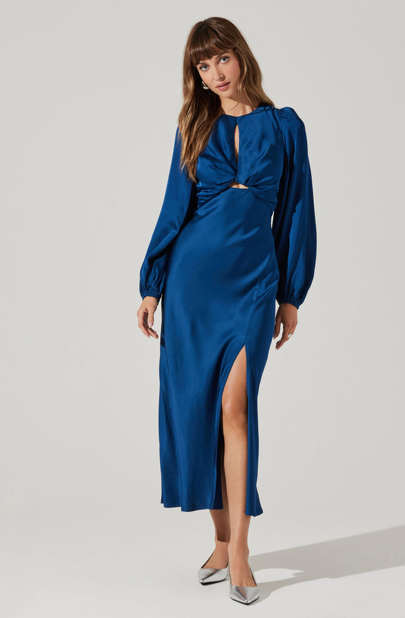 A woman is wearing a long, royal blue satin dress with long, puffed sleeves gathered at the cuffs. The dress features a keyhole neckline with a twist detail at the front, creating a small cutout at the bust. It has a high slit on one side, revealing her leg. She pairs the dress with pointed silver high heels.