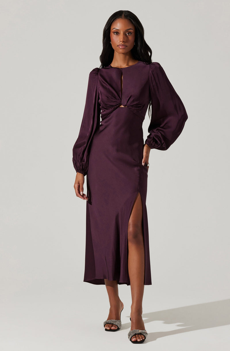 A woman is wearing a long, dark purple satin dress with long, puffed sleeves. The dress features a cut-out detail at the waist and a high slit on one side, revealing her leg. She is also wearing a pair of open-toe high heels with embellishments.