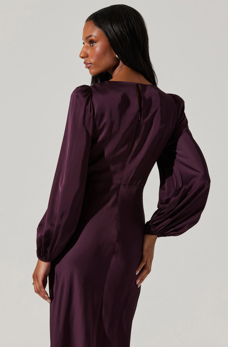 The dress is a deep, rich purple satin fabric with a subtle sheen. It features long, voluminous puff sleeves that are gathered at the wrists with elastic. The dress has a fitted waist, and the back has a small keyhole opening at the neckline. The material appears smooth and elegant, giving it a formal and sophisticated look.