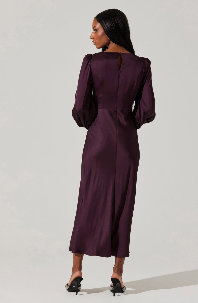 A woman is wearing a long, elegant dress made of shiny, deep purple satin fabric. The dress features puffed long sleeves with gathers at the shoulders, a high neckline, and a back zipper closure. The skirt is fitted at the waist and flows down to her ankles, with a slit at the back. She is also wearing black high-heeled sandals with ankle straps.