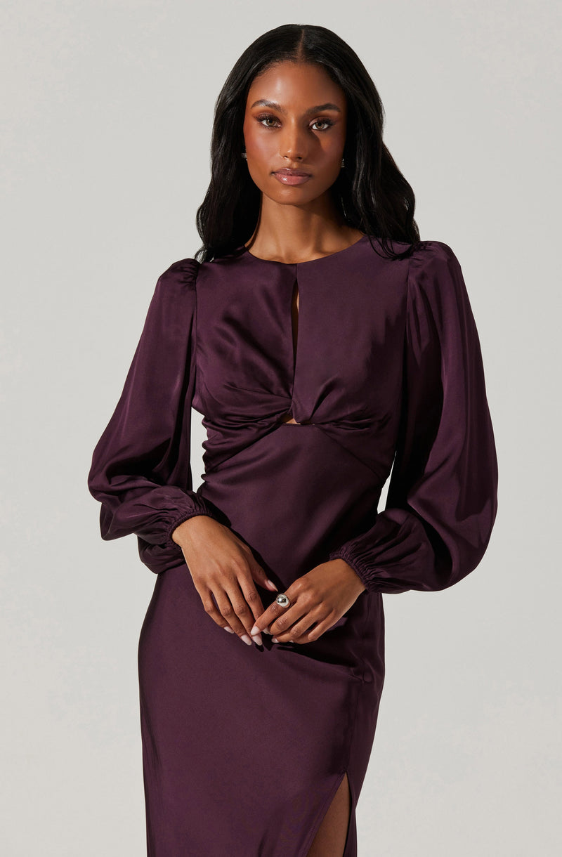 A woman is wearing a deep purple satin dress with long, puffed sleeves gathered at the wrists. The dress has a round neckline with a small keyhole opening at the front and features gathered fabric at the bust. The skirt is fitted with a subtle slit on one side.