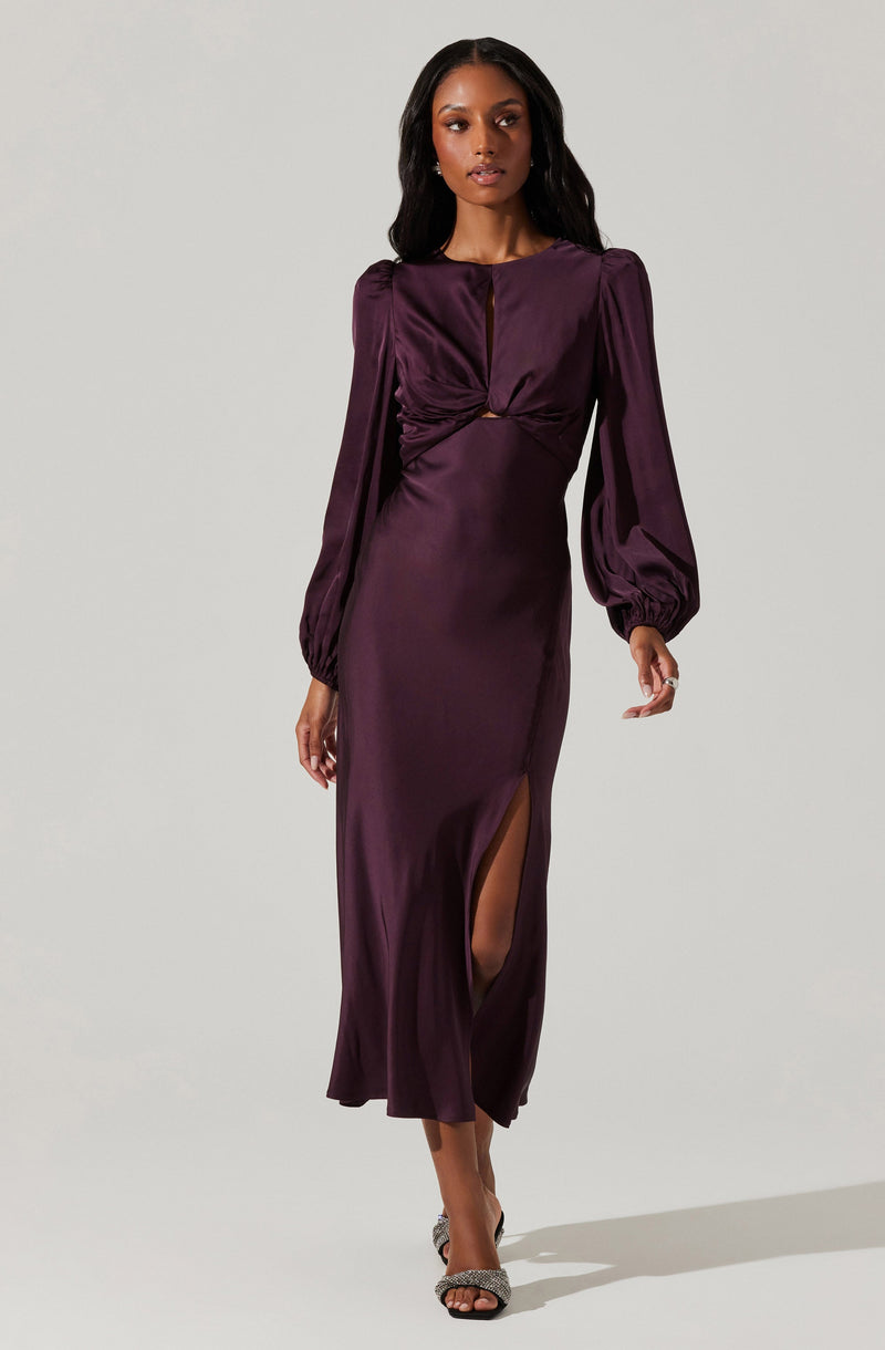 A woman is wearing a long, deep purple satin dress with long puffed sleeves gathered at the cuffs. The dress features a cut-out detail at the waist and a high slit on one side. She is also wearing high-heeled sandals with embellishments.