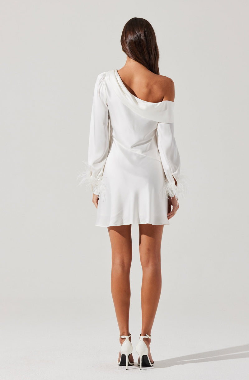 A woman is wearing a white satin dress with an off-the-shoulder neckline on the right side, exposing her shoulder and collarbone. The dress has long sleeves that gather at the wrists with feathered cuffs, and it has a slightly flared skirt that falls above the knee. She is also wearing white high-heeled shoes with ankle straps.