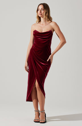 A woman is wearing a strapless, deep burgundy velvet dress with a fitted bodice and side slits that reveal her legs. The dress features gathered and draped fabric details across the front. She pairs the dress with black high-heeled sandals with ankle straps.
