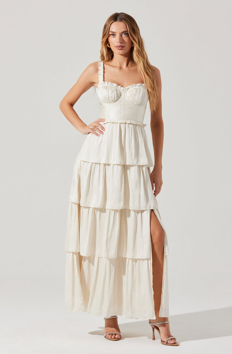 A woman is wearing a cream-colored, sleeveless maxi dress with ruffled shoulder straps. The dress has a fitted bodice with subtle textured detailing and a tiered, flowing skirt with a high slit on one side. She is also wearing nude high-heeled sandals.