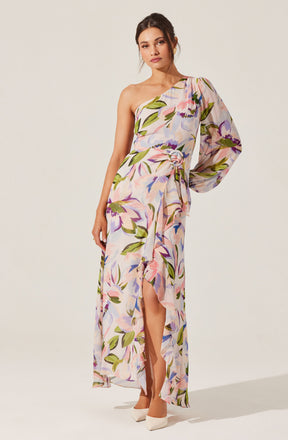 A woman is wearing a long, flowing dress with a floral pattern. The dress features a prominent one-shoulder design with a puffed sleeve on her right side and sleeveless on her left. The fabric has a pastel-colored floral print with shades of purple, pink, green, and blue. The dress has a slit up the front, revealing her legs and white high-heeled shoes. There is a matching fabric belt tied at the waist.