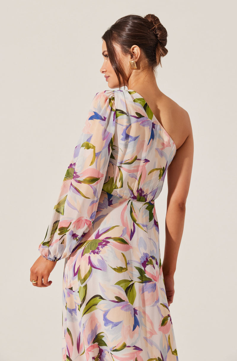 A woman is wearing a flowy, floral dress with a one-shoulder design. The dress features large, colorful flowers in shades of pink, purple, blue, and green on a light background. The dress has a long sleeve on one side with a puffed shoulder, and the other side is sleeveless. The fabric appears lightweight and semi-sheer.