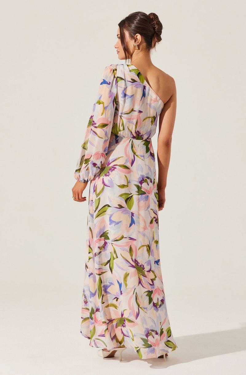 A woman is wearing a long, flowing dress with a floral pattern featuring large pink, purple, and blue flowers with green leaves. The dress has one long sleeve on the left side and is sleeveless on the right side, with a high waistline and a slightly cinched waist.