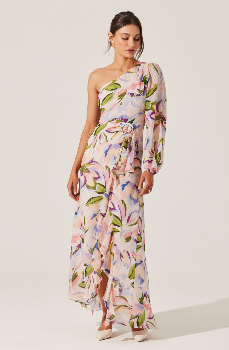 A woman is wearing a long, flowy dress with a pastel-colored floral print. The dress features an asymmetrical design with a one-shoulder style, including a long sleeve on one side and sleeveless on the other. The fabric is lightweight and semi-transparent, and the dress has a ruffled hem with a slit. She is also wearing nude high-heeled shoes.