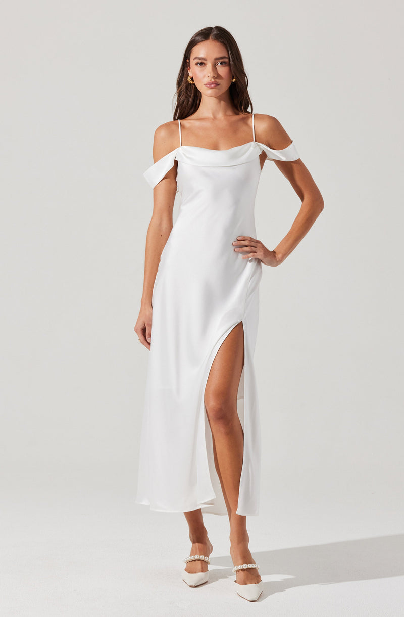 A woman is wearing a white, off-the-shoulder dress with spaghetti straps. The dress features ruffled details on the shoulders and a thigh-high slit on one side. She is also wearing white heels with pearl embellishments.