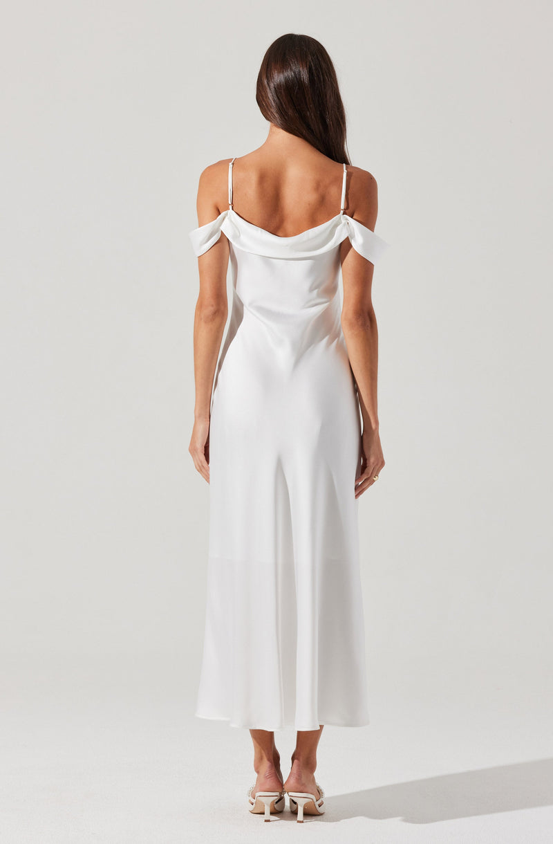 A woman is wearing a white, off-the-shoulder dress with spaghetti straps. The dress has a draped neckline with bow details on the shoulders and flows down to a wide-leg length. She is also wearing white high-heeled shoes with a bow detail.