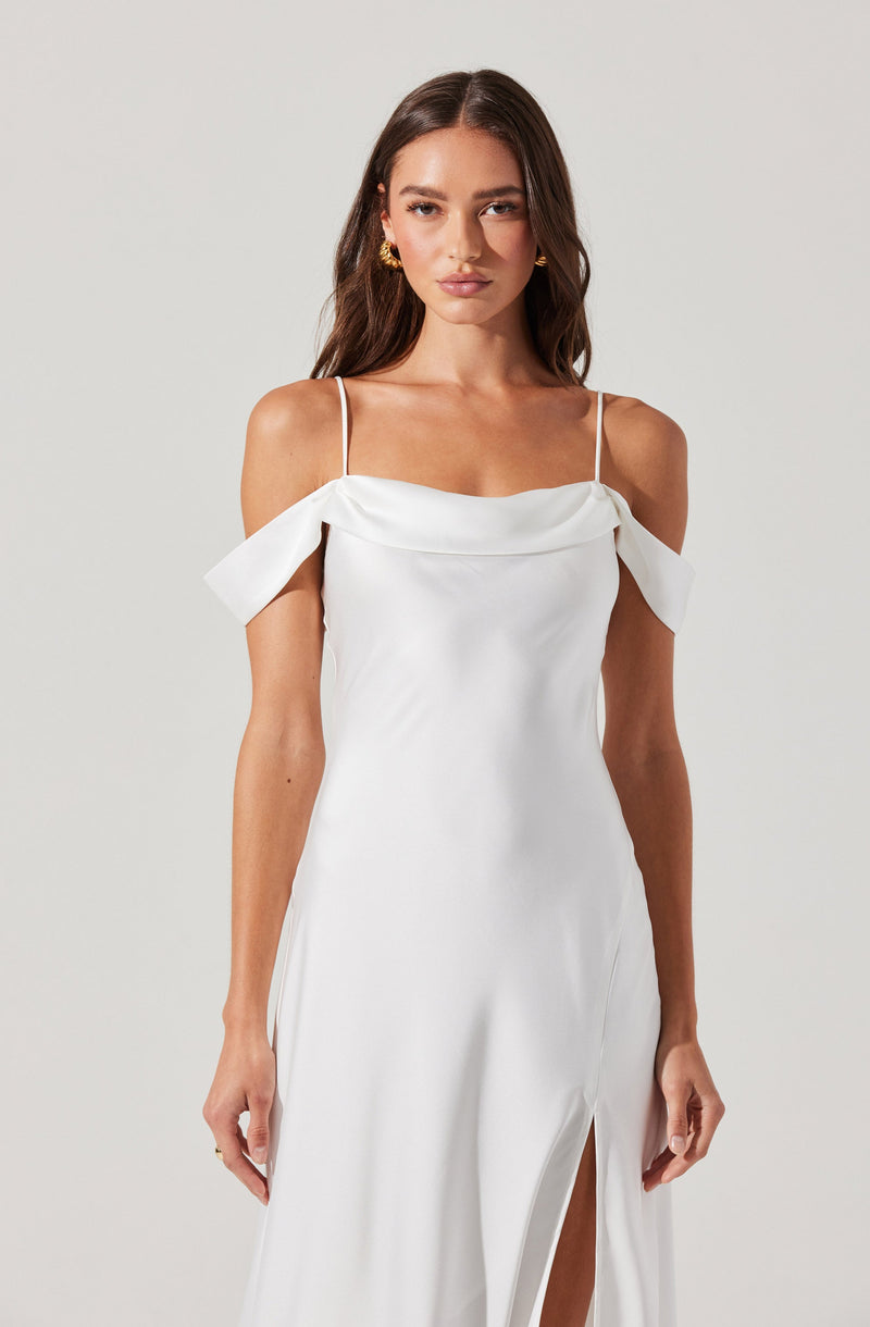 A woman is wearing a white gown with thin spaghetti straps. The dress features off-the-shoulder ruffled sleeves and a high slit in the front, revealing her legs.