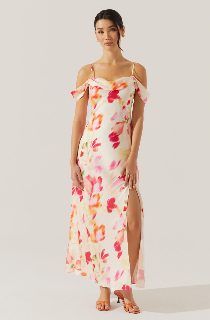 A woman wearing a long, flowy dress with a floral pattern featuring shades of pink, red, orange, and yellow. The dress has thin spaghetti straps, off-the-shoulder flutter sleeves, and a high slit on one side.