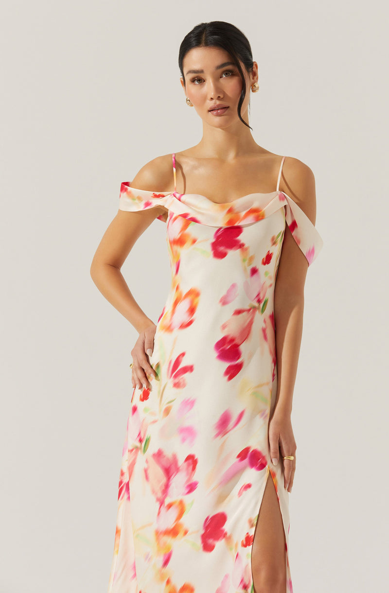 A woman is wearing a sleeveless, floor-length dress with a floral pattern in shades of pink, red, orange, and green on a light cream background. The dress features a draped off-the-shoulder neckline with thin spaghetti straps and a high slit on the side.
