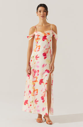 A woman is wearing a long, sleeveless, off-the-shoulder dress with spaghetti straps. The dress features a watercolor floral pattern in shades of pink, red, orange, and green on a white background. It has a slit on one side, revealing her leg. She is also wearing open-toe heels.