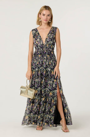 A woman is wearing a sleeveless, maxi-length dress with a deep V neckline. The dress has a black background with a colorful floral pattern including purple, yellow, and green flowers. It features a cinched waist and a high slit on one side, revealing her leg. She is accessorizing with large, round pearl earrings and is carrying a small, light-colored handbag. She is also wearing strappy, open-toe heels.