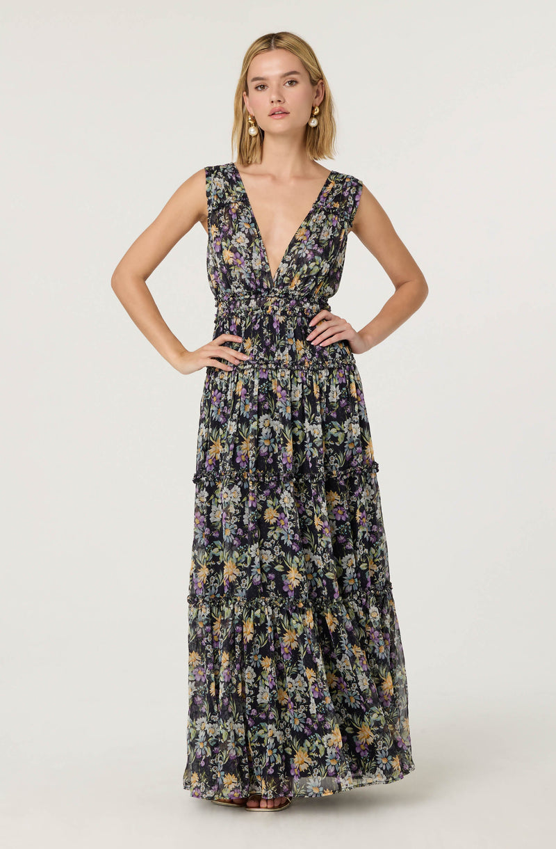 The dress features a sleeveless design with wide shoulder straps and a deep V-neckline. It has a fitted waist and a long, flowing skirt divided into three tiers, each adorned with a vibrant floral pattern. The fabric appears lightweight and semi-sheer, suited for warm weather.