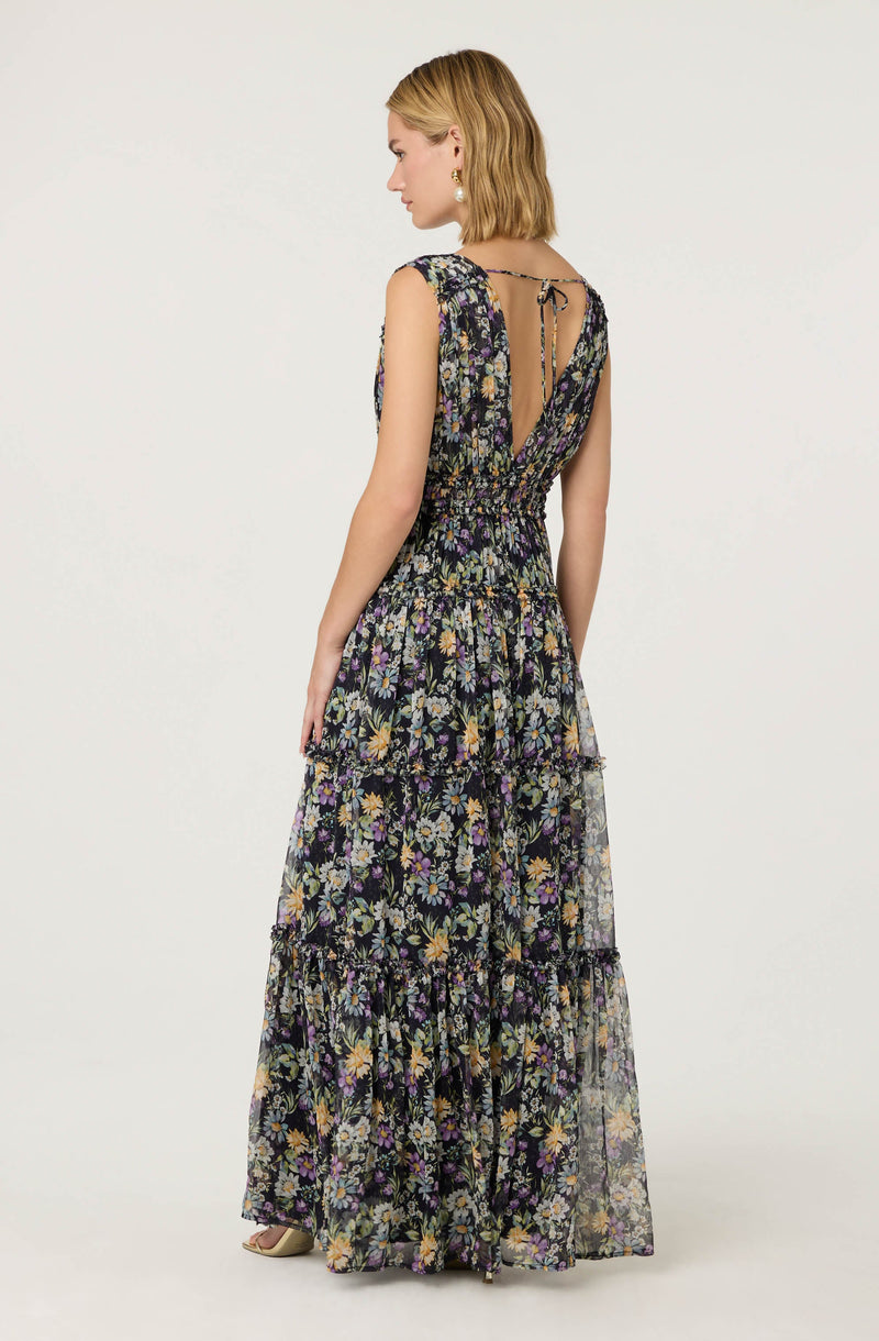 A woman is wearing a sleeveless, floor-length dress with a floral pattern. The dress has a fitted waist with gathered fabric and a tiered skirt. The fabric features various flowers in shades of purple, yellow, and green on a dark background. The back of the dress has a deep V opening with a lace-up tie at the top.