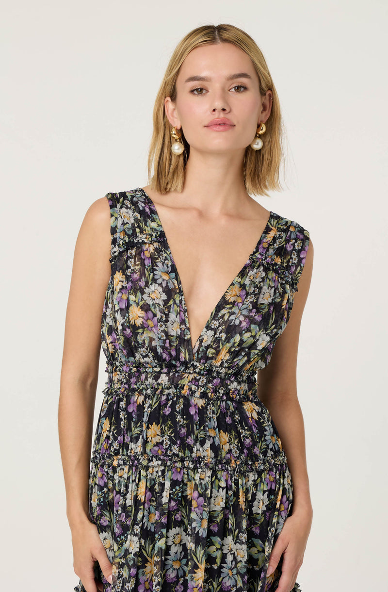 The woman is wearing a sleeveless dress with a deep V neckline and an elastic waistband. The dress features a floral pattern with various flowers in shades of purple, yellow, white, and green on a dark background. The fabric appears lightweight and flowy.
