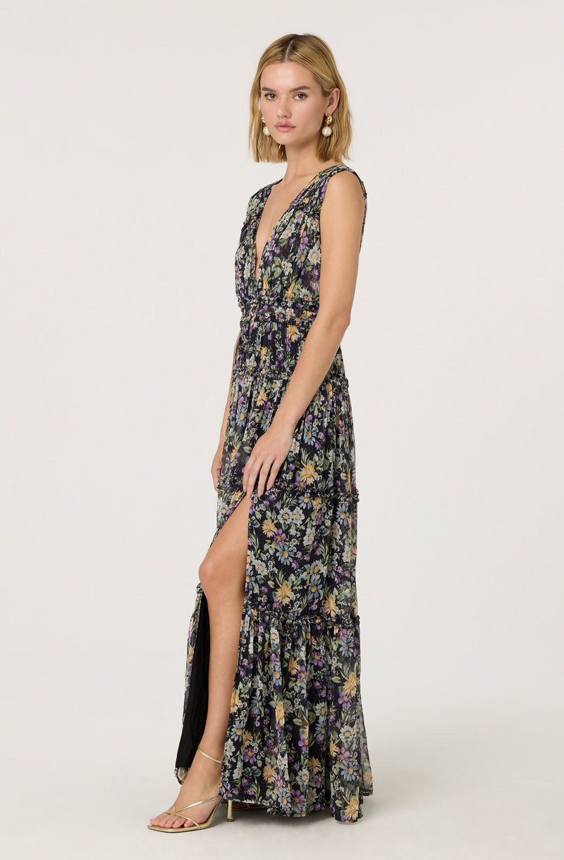 A woman is wearing a sleeveless, long floral dress with a deep V neckline. The dress features a multicolored floral pattern on a dark background, with layers and a high slit revealing one leg. She is accessorized with large pearl earrings and strappy high-heeled sandals.