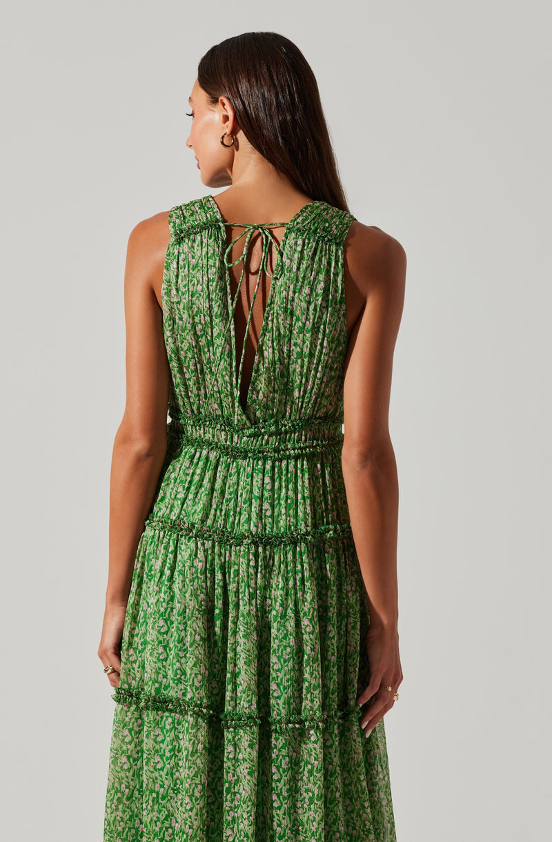 A woman is wearing a sleeveless, green dress with a small, intricate pattern. The dress features a deep V-back with a tie closure at the top, and the fabric is gathered at the waist, creating a slightly cinched look. The skirt is tiered, with horizontal seams separating each gathered section, giving the dress a flowing, layered appearance.