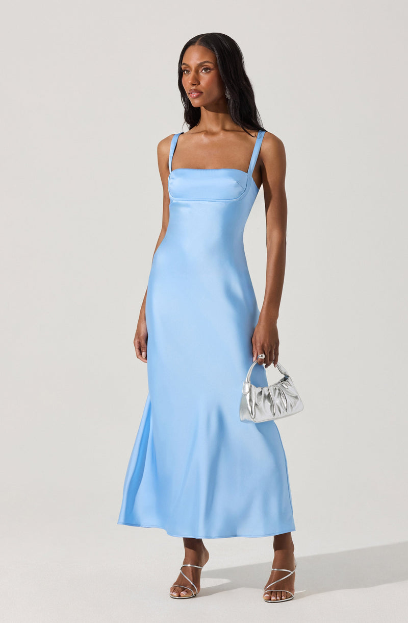 A woman is wearing a light blue, floor-length satin dress with a fitted bodice and thin shoulder straps. She is accessorizing with a small silver handbag and strappy high-heeled sandals.